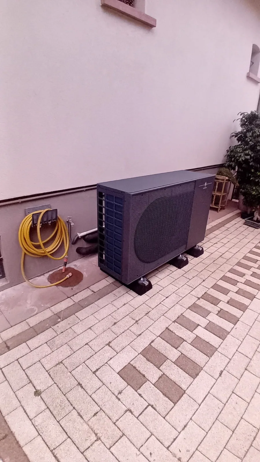 Groupe extérieur de la pompe à chaleur air-eau sur plots, vu depuis l'allée pavée, chez notre client à Achenheim (Bas-Rhin).